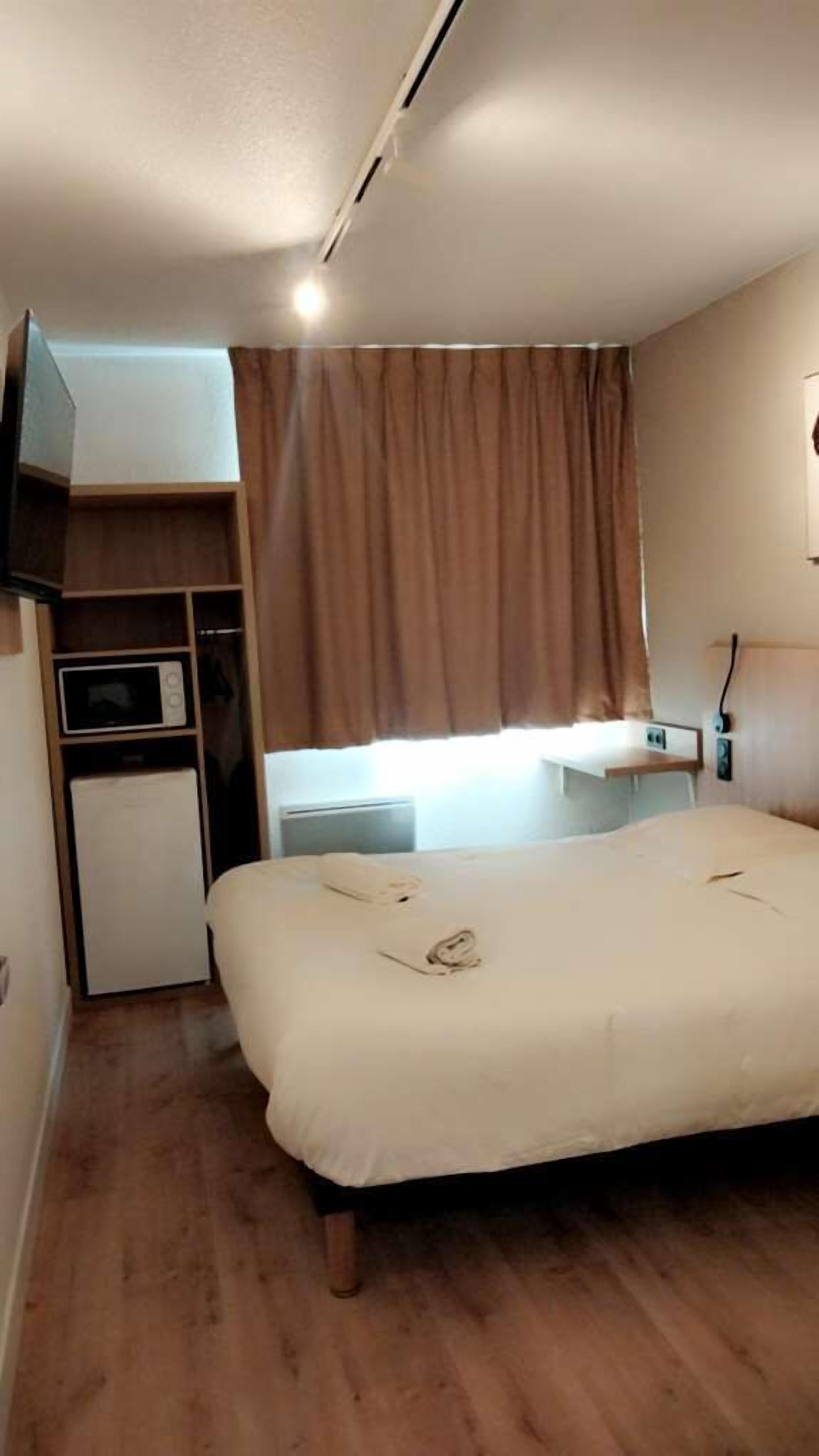 A bedroom with a bed, a built-in TV, a small kitchenette, and a window with curtains.