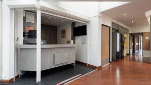 Zona de recepción de hotel con mostrador de recepción, mesa de seguridad, televisor montado en la pared y un pasillo que conduce a otras habitaciones.