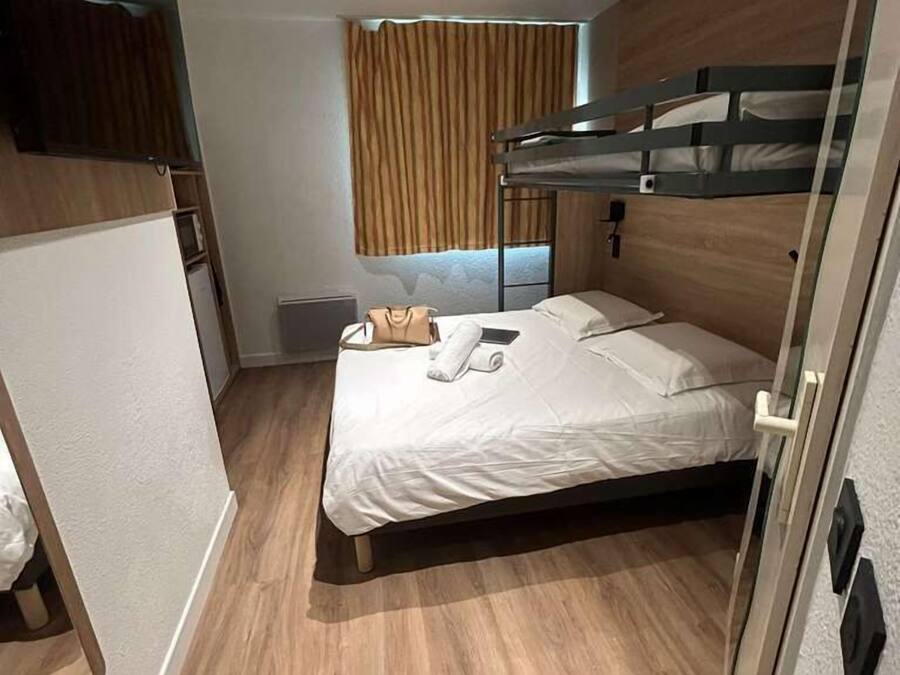 A compact hotel room with a bunk bed, wooden flooring, a small bedside table, and a window with curtains.