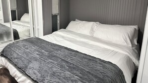 Un llit ben arrenjat amb llençols blancs i una manta gris, una finestra amb persianes, i un mirall que reflecteix part de l'habitació.