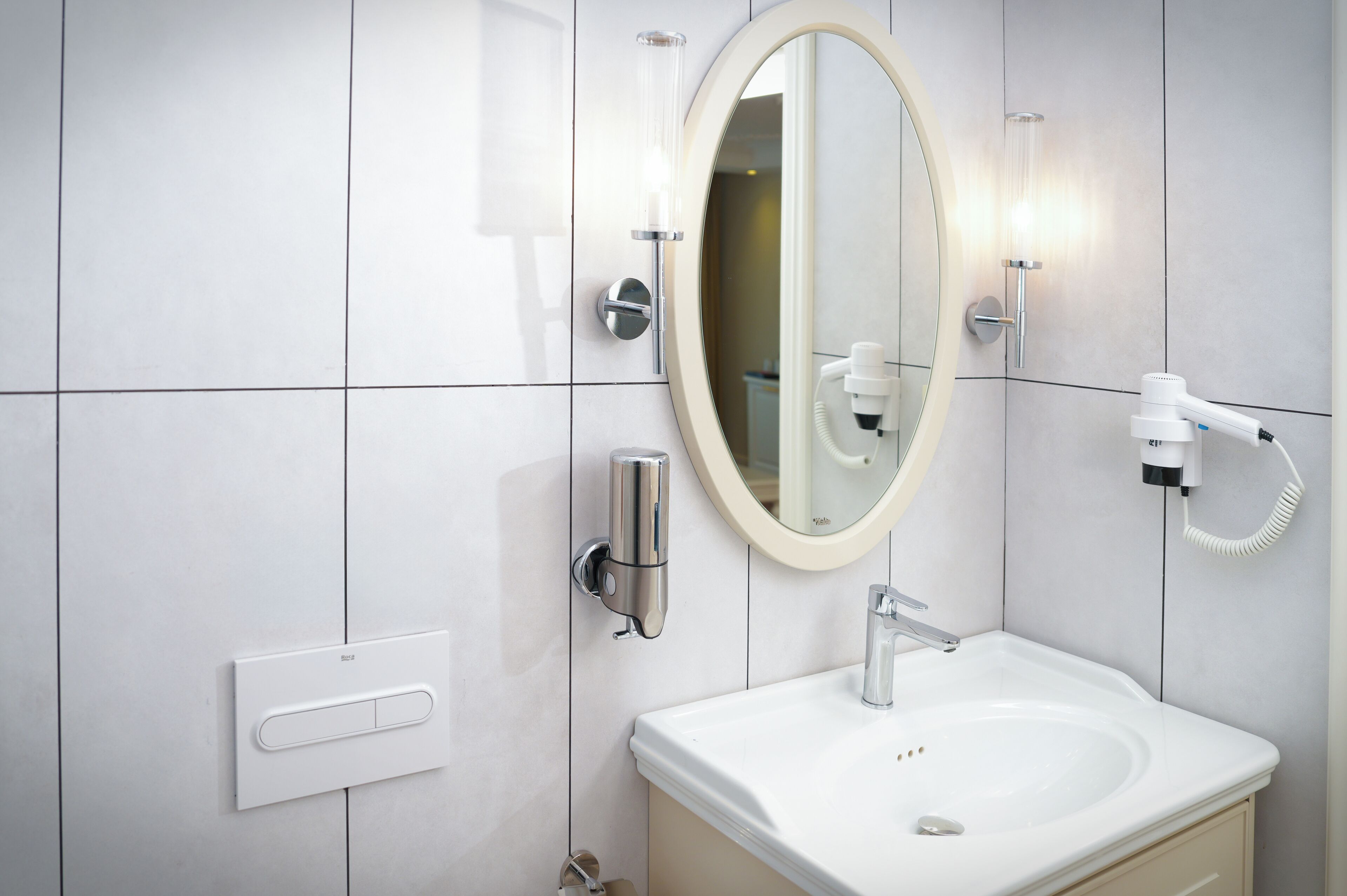 Un baño moderno con un lavamanos blanco, un espejo redondo, un cabezal de ducha manual y un dispensador de jabón de pared.