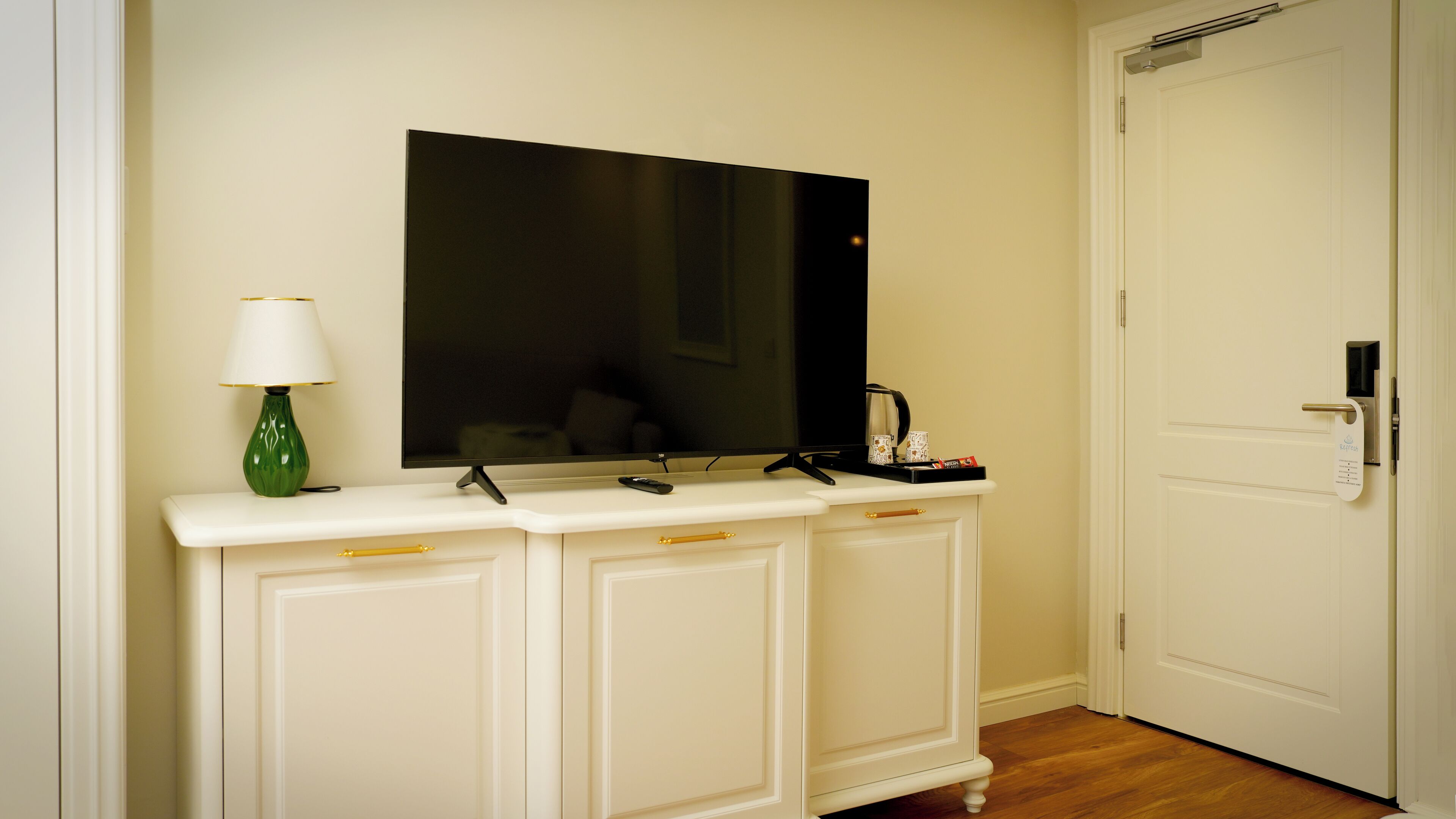 A white console table with a flat-screen TV, a green vase, and a tray with a remote control and a mug.
