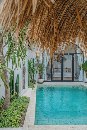 A pool area with a thatched roof, surrounded by potted plants and a glass-enclosed bedroom. - Luana Villa by Betterplace (Canggu)