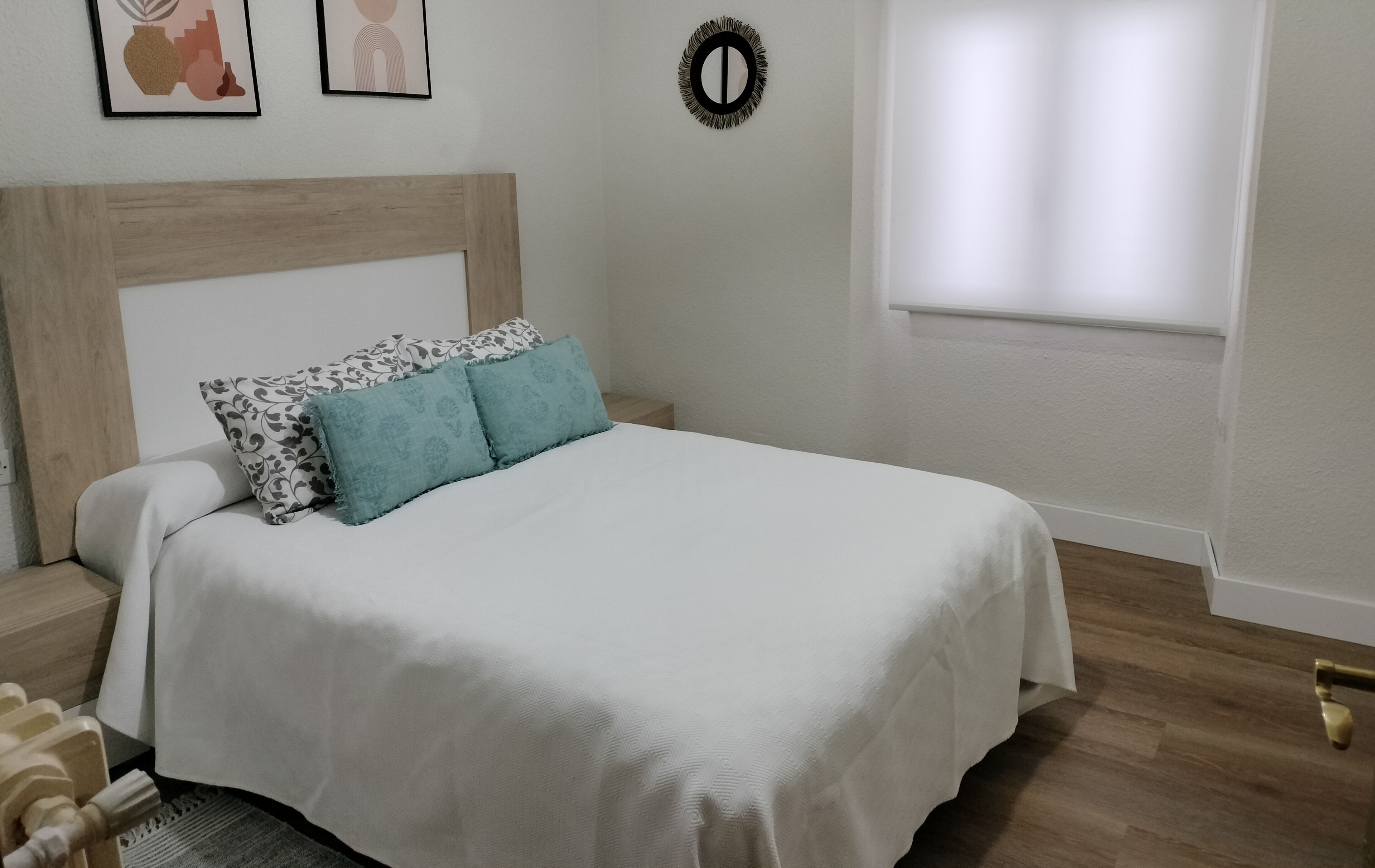 Una cama bien hecha con edredón blanco y almohadas estampadas, cabecera de madera y mesita de noche con lámpara.