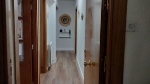 A narrow hallway with wooden flooring and white walls. There is a door on the right and a radiator on the left. A shelf with decorative items and a mirror on the wall is visible in the background.