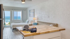 Ein Hotelzimmer mit Bett, einem Holztisch, einem Deckenventilator, Blick auf den Strand und einem Bild an der Wand.