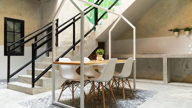 A modern interior with a dining area featuring a table and chairs, a staircase with a black railing, and a concrete wall with potted plants. - Fybed Guesthouse (Semarang)