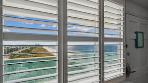 A room with a large window featuring horizontal blinds, a view of a coastal area with a beach and ocean, and a door with a green-framed sign. - Fontainebleau 706 by AvantStay | Beachfront+Views (Panama City Beach)