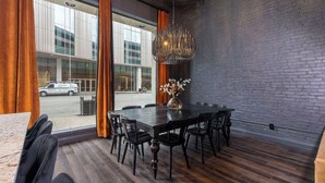 A modern dining area with a dark wooden table, black chairs, and a decorative metal chandelier. Large windows offer a view of the street outside. - Historic District Hideaway. 4bed (Louisville)