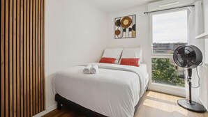 A bedroom with a bed, a wall-mounted artwork, a window with a view, and a standing fan. - Chic and comfortable apart with Parking and balcony (Bezons)