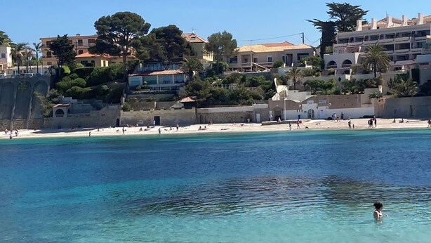 Uma praia costeira com águas turquesa cristalinas, areal arenoso e edifícios ao fundo.