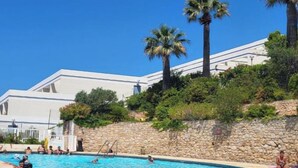 Uma piscina com pessoas, cercada por um muro de pedra e palmeiras, com um edifício ao fundo.