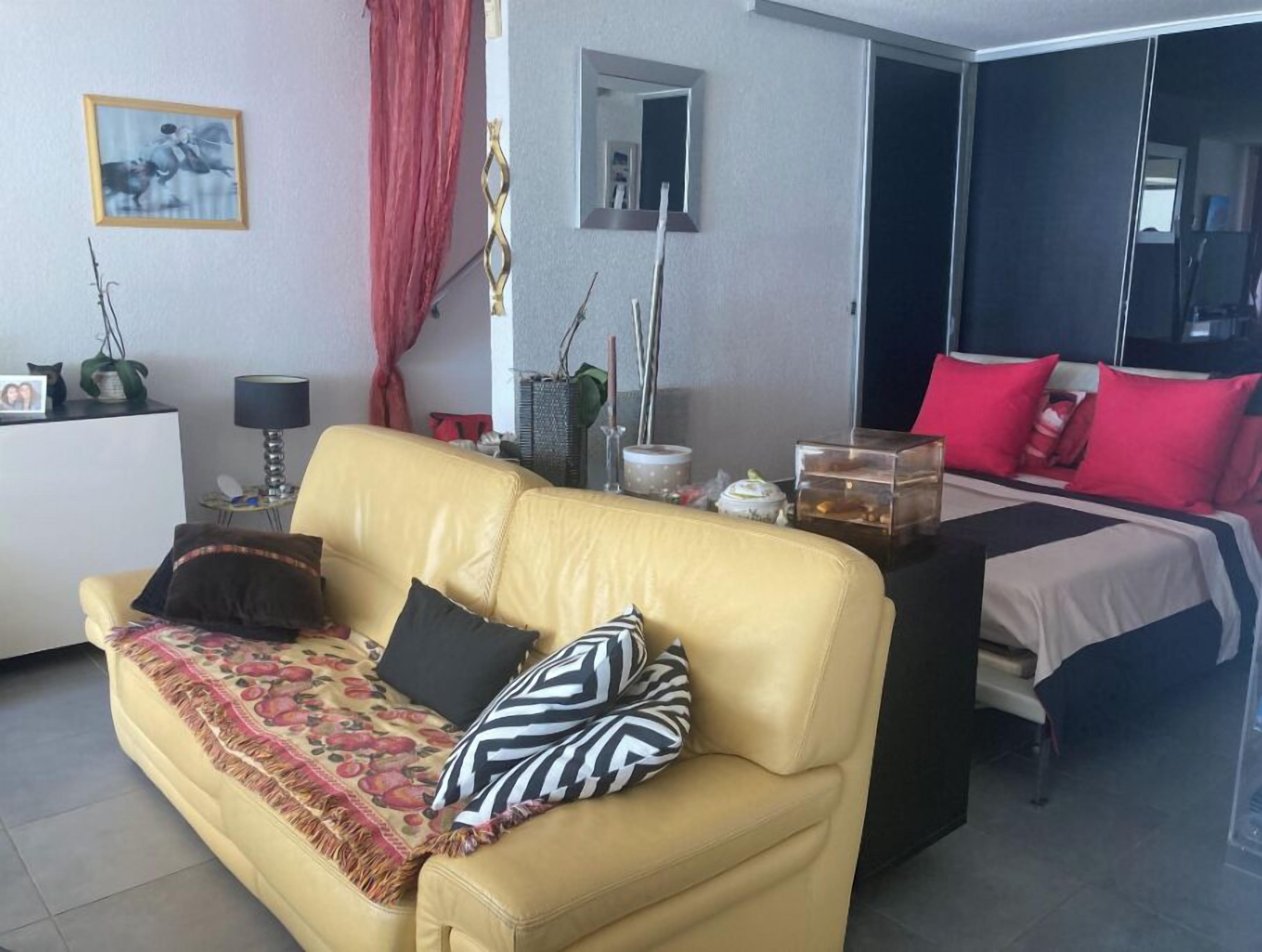 A modern living room with a yellow sofa, a bed with red pillows, a small table with decorative items, and a mirror on the wall.
