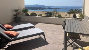 Uma varanda com duas cadeiras de lounge e uma mesa, com vista para o mar e montanhas ao longe.