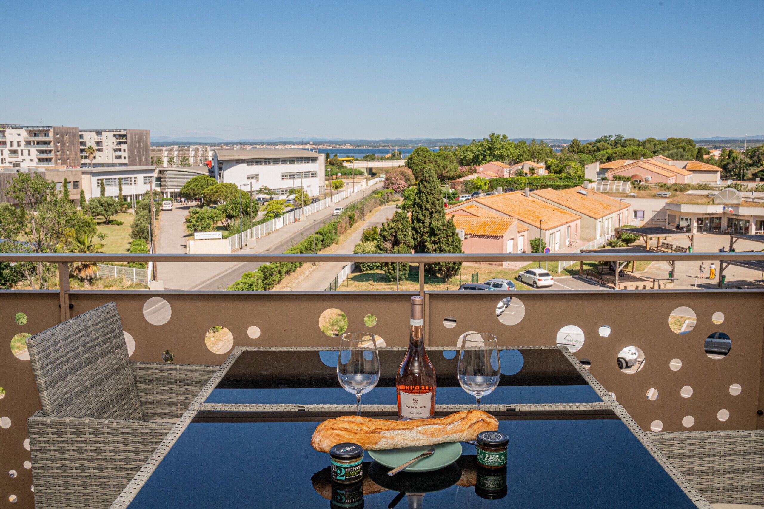 A balcony with a table set for two, a bottle of wine, a baguette, and two cans of beer.