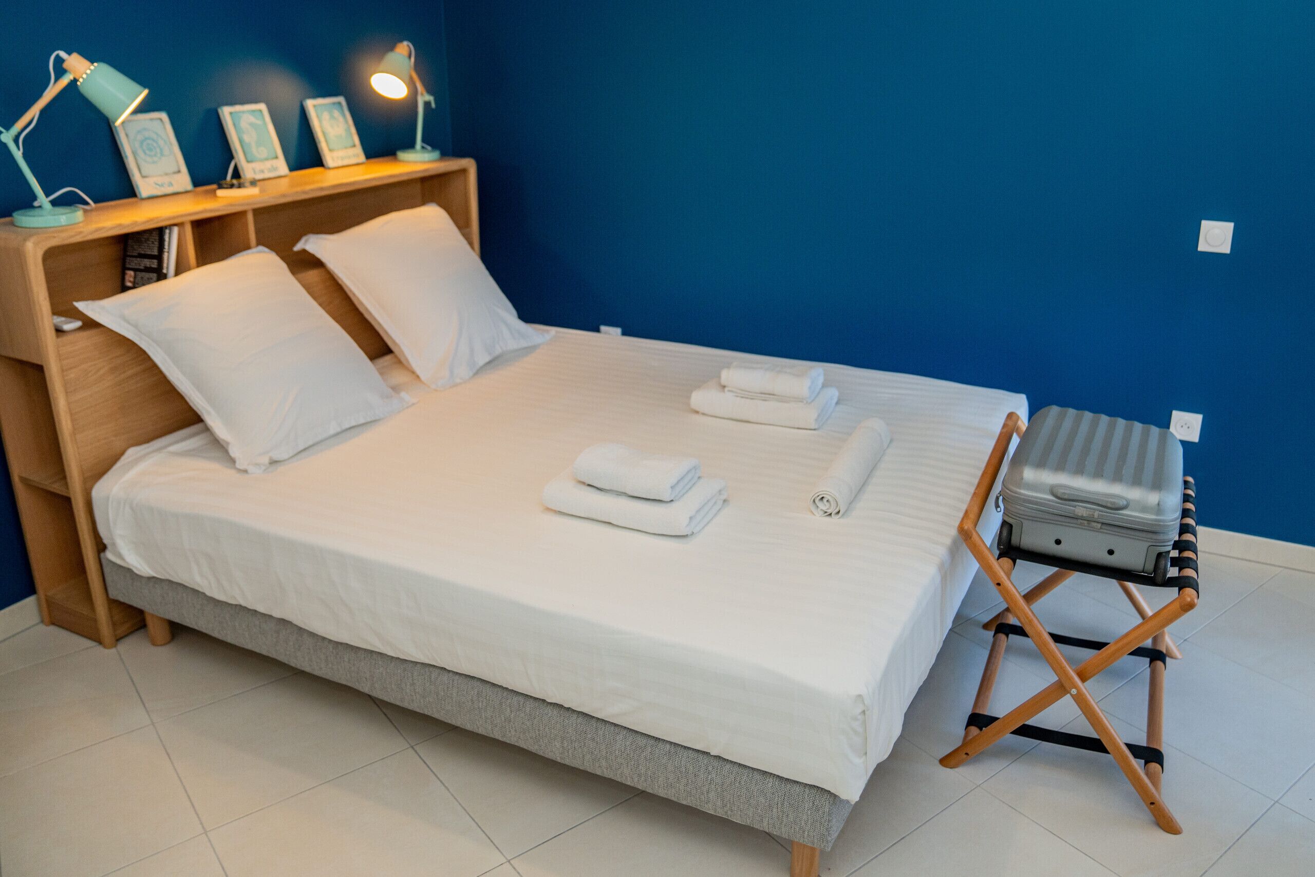 Un lit bien fait, avec du linge de lit blanc, une tête de lit en bois et une petite table de chevet sur laquelle se trouve une lampe.