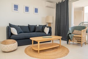 A modern living room with a dark blue sofa, a wooden coffee table, a woven pouf, and a wicker chair. - Sète, Apt 2 chambres, Terrasse et proximité plages (Sète)