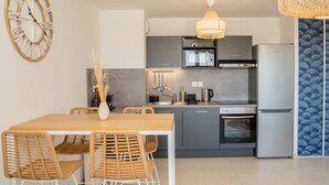 Eine moderne Küche mit grauen Schränken, einem Edelstahlkühlschrank, einer Mikrowelle und einem Backofen. Ein Holztisch mit Stühlen und eine große Wanduhr sind ebenfalls vorhanden.