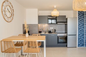 A modern kitchen with grey cabinets, a stainless steel refrigerator, a microwave, and an oven. There is a wooden dining table with chairs and a large wall clock. - Sète, Apt 2 chambres, Terrasse et proximité plages (Sète)