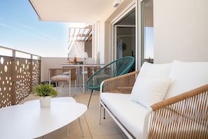 A balcony with a white sofa, a green chair, a small table with a potted plant, and a dining area with chairs and a table.