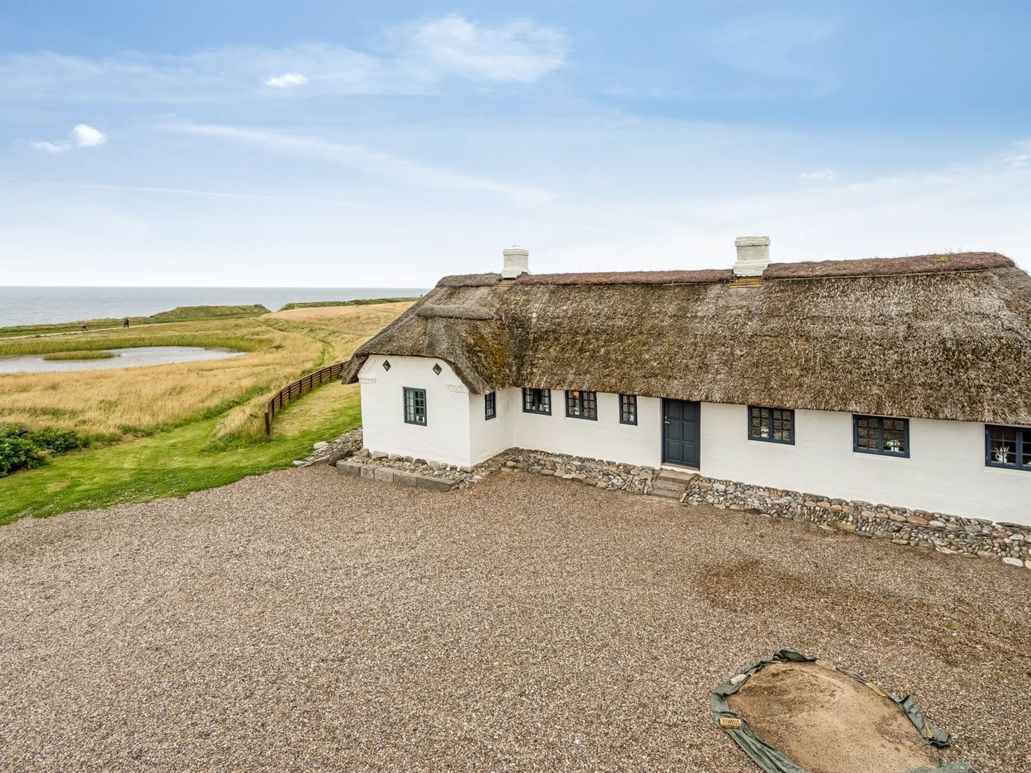 A white house with a thatched roof, situated on a gravel driveway with a view of the sea and grassy fields.