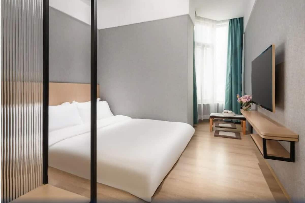 Ein modernes Hotelzimmer mit einem großen Bett, einem Nachttisch, einem an der Wand montierten Fernseher und einem Fenster mit Vorhängen.