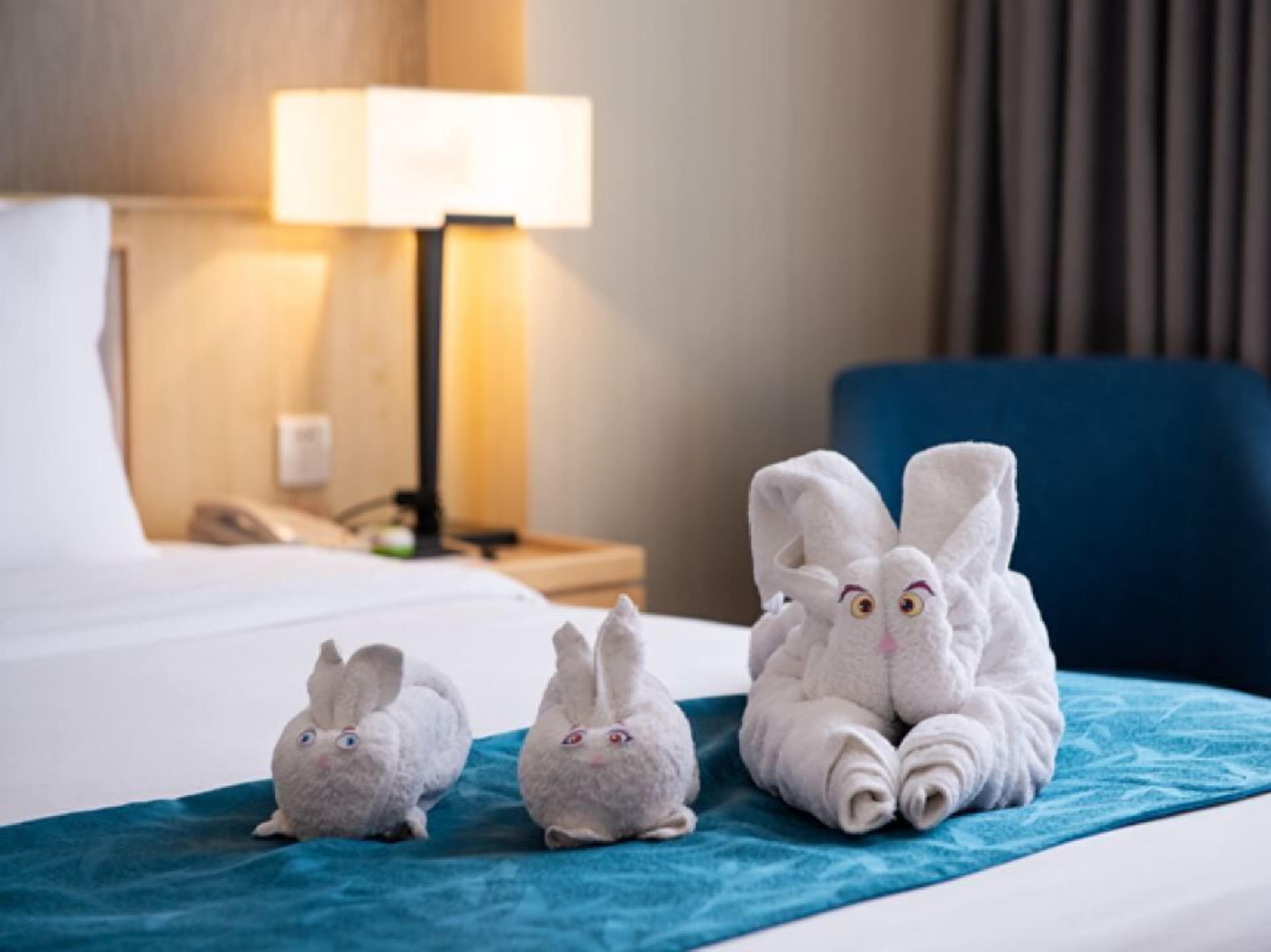 Quarto de hotel com uma cama, uma mesa de cabeceira com abajur e três toalhas macias em formato de coelho na cama.