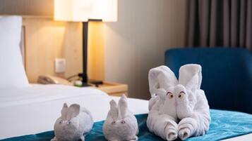 Quarto de hotel com uma cama, uma mesa de cabeceira com abajur e três toalhas macias em formato de coelho na cama.