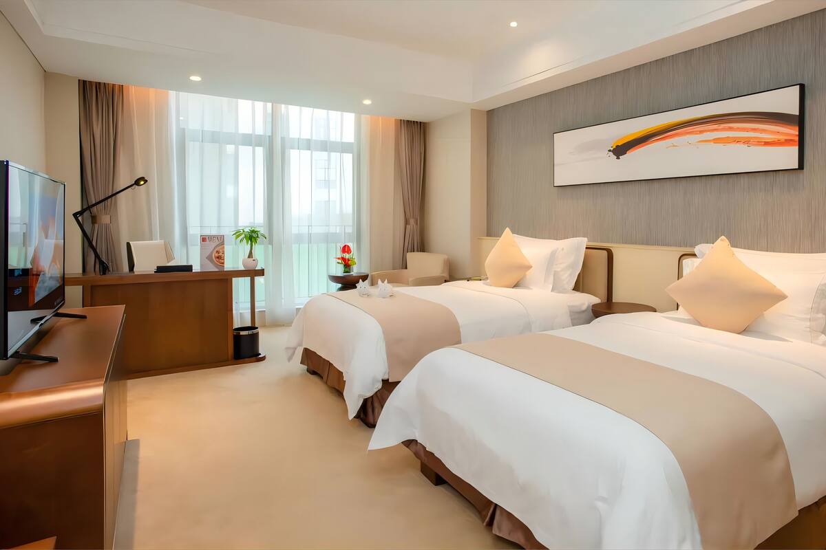 A hotel room with two beds, a desk with a computer, a TV, and a large window with curtains.