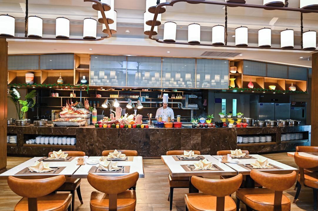 En restaurant med en kok ved disken, borde dækket med hvide duge og brune stole, samt et moderne interiør med hængende lamper.