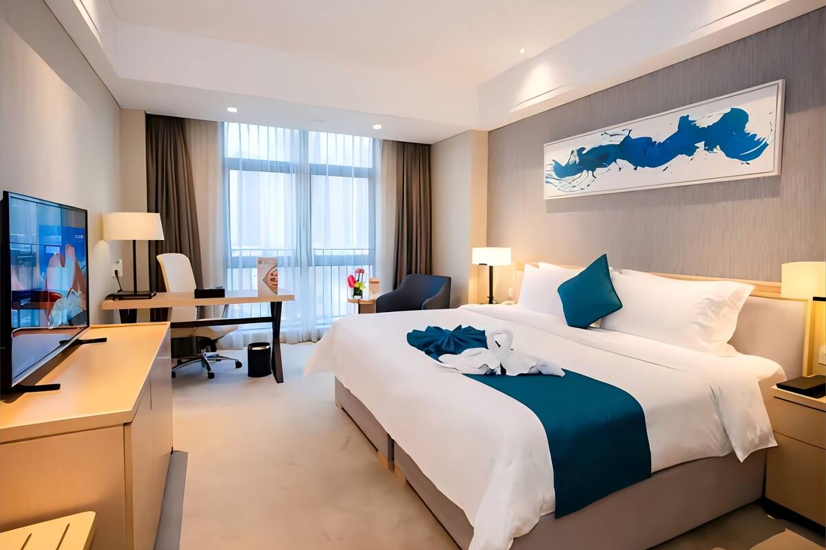 A modern hotel room with a large bed, a desk with a TV, and a painting on the wall.