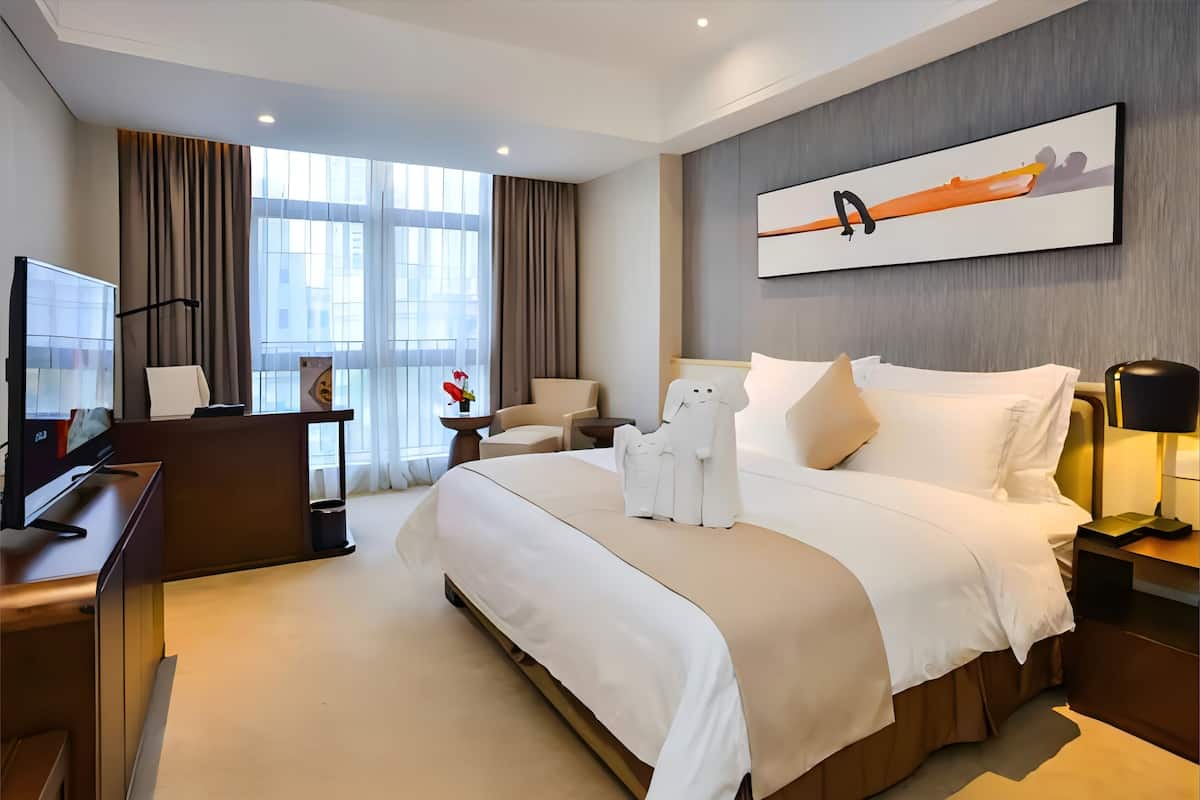 A hotel room with a large bed, a desk with a computer, a chair, a small table with a vase, and a painting on the wall.
