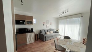 A compact living space with a kitchenette, a grey sofa, a dining table with chairs, and a window with sheer curtains. - Appartement Cosy au Centre de Meaux (Meaux)