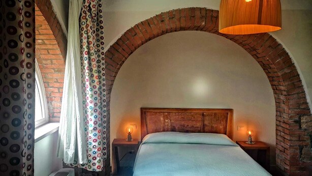 Un dormitorio con techo abovedado de ladrillo, una cama con cabecera de madera, dos lámparas de noche y una ventana con cortinas estampadas.