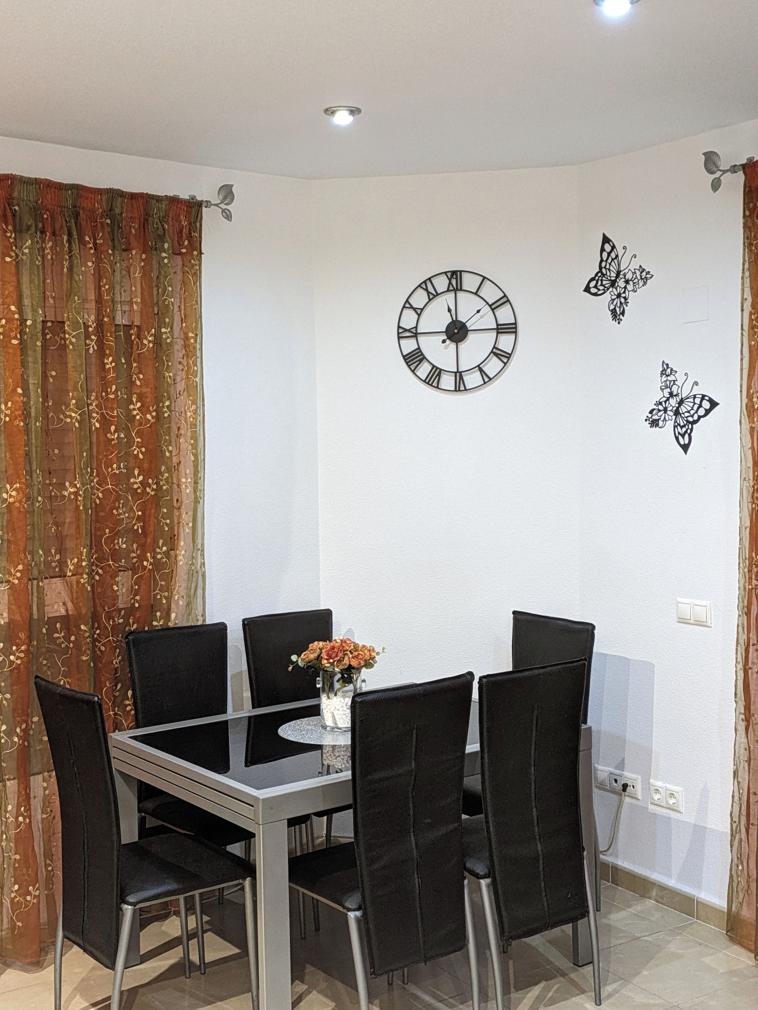 Un comedor con una mesa preparada para cuatro personas, rodeada de sillas, un reloj de pared y decoración de mariposas en la pared.