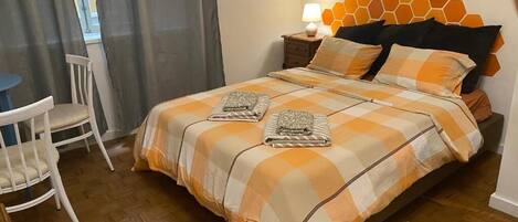A bedroom with a checkered bedspread, a bedside lamp, a chair, and a window with curtains.
