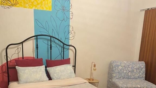 A bed with a red and white bedspread, a metal frame, and two pillows. There is a chair with a patterned cushion and a small table with a lamp. The room has a wooden floor and a rug. - Barão 35 Guest House (Braga)