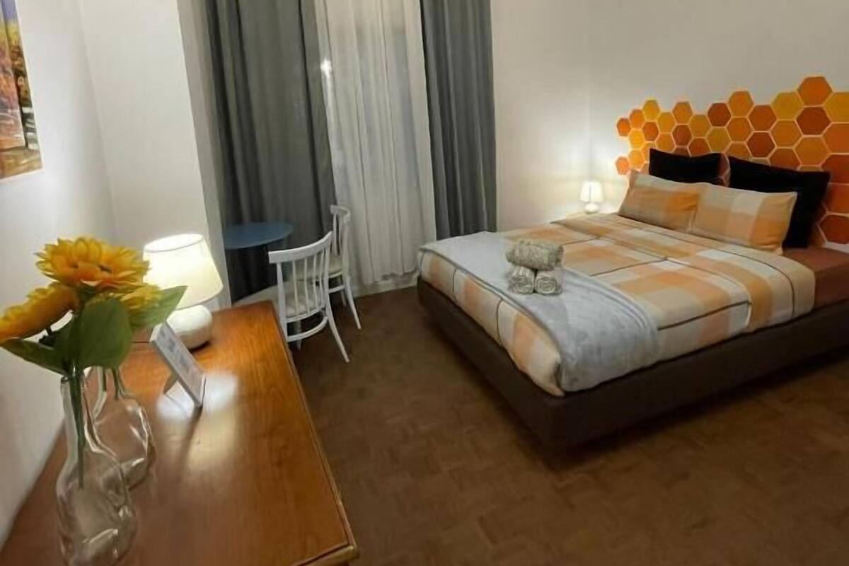 Une chambre d’hôtel avec un lit, un bureau, une chaise, un vase de fleurs et une fenêtre avec des rideaux.