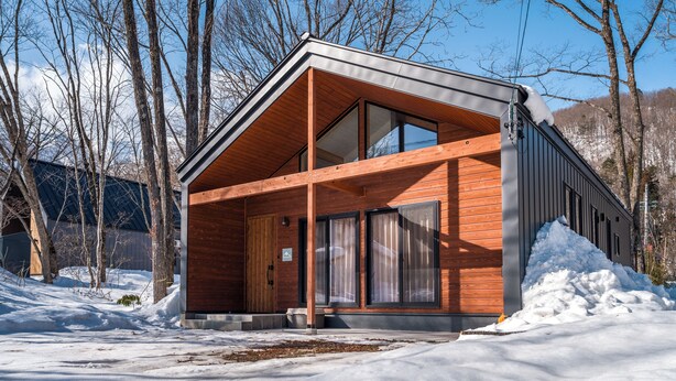 Una cabaña moderna de madera con amplios ventanales, techo inclinado y suelo cubierto de nieve.