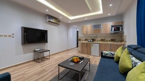 Ein modernes Wohnzimmer mit Fernseher, einem kleinen Tisch mit Früchten, einer Couch mit Kissen und einer Küchenzeile mit Schränken und Geräten.