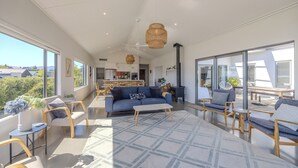 A spacious living area with a blue sofa, wooden chairs, a white coffee table, and large windows offering a view of the outdoors. - Lake View Heights - Luxury large modern House (244 sq mtr)  (Wanaka)