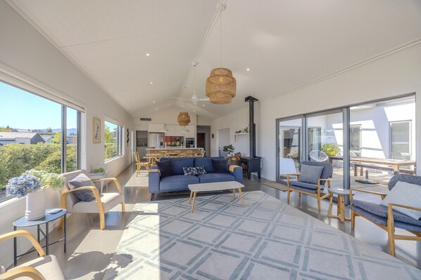 A spacious living area with a blue sofa, wooden chairs, a white coffee table, and large windows offering a view of the outdoors. - Lake View Heights - Luxury large modern House (244 sq mtr) (Wanaka)