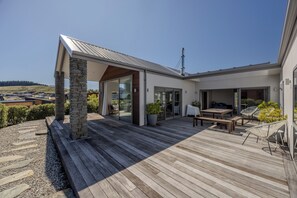 A modern outdoor deck with a stone column, a wooden pergola, and a seating area with chairs and a table. - Lake View Heights - Luxury large modern House (244 sq mtr) (Wanaka)