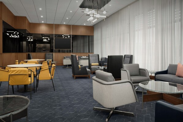 Un vestíbulo moderno de hotel con una zona de estar central, un bar y un ventanal con cortinas translúcidas.