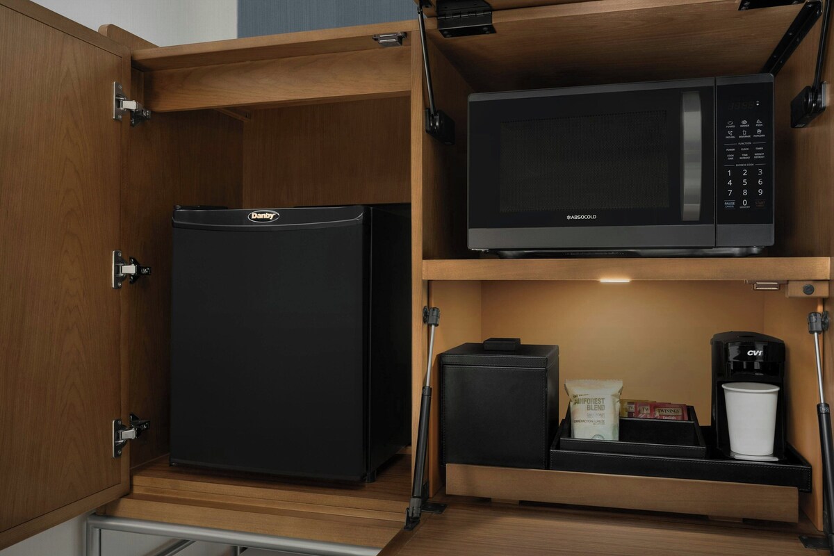 Cocina pequeña con un refrigerador negro, un microondas, una cafetera y un mueble bajo con una taza de café y algunos objetos.