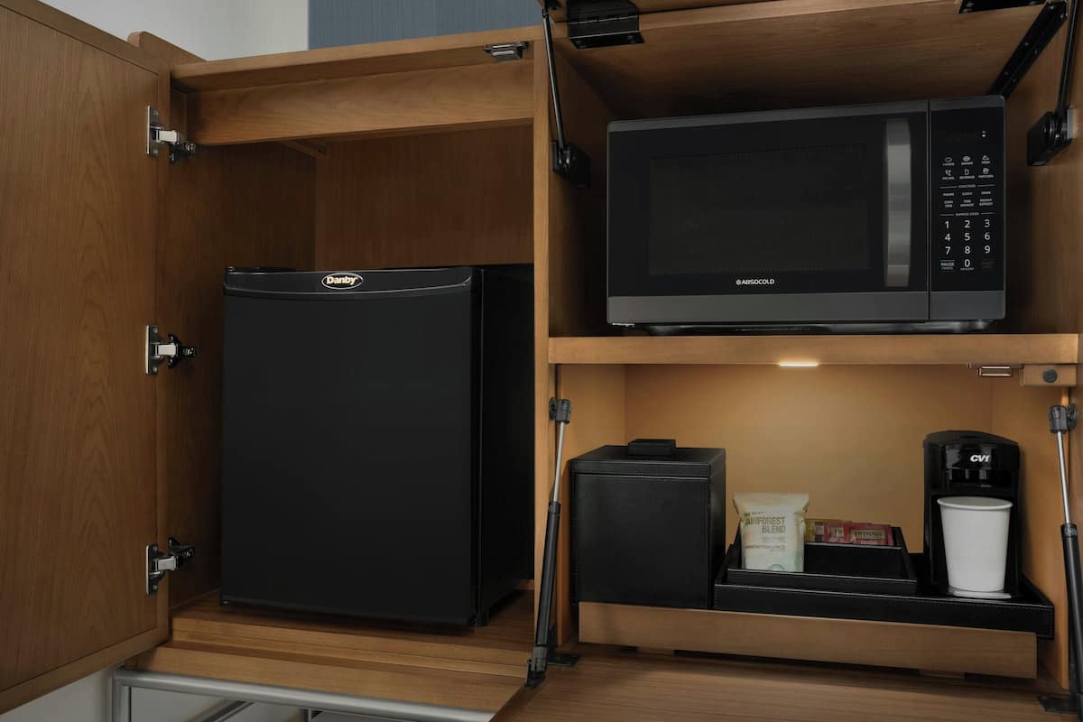 Cocina pequeña con un refrigerador negro, un microondas, una cafetera y un mueble bajo con una taza de café y algunos objetos.