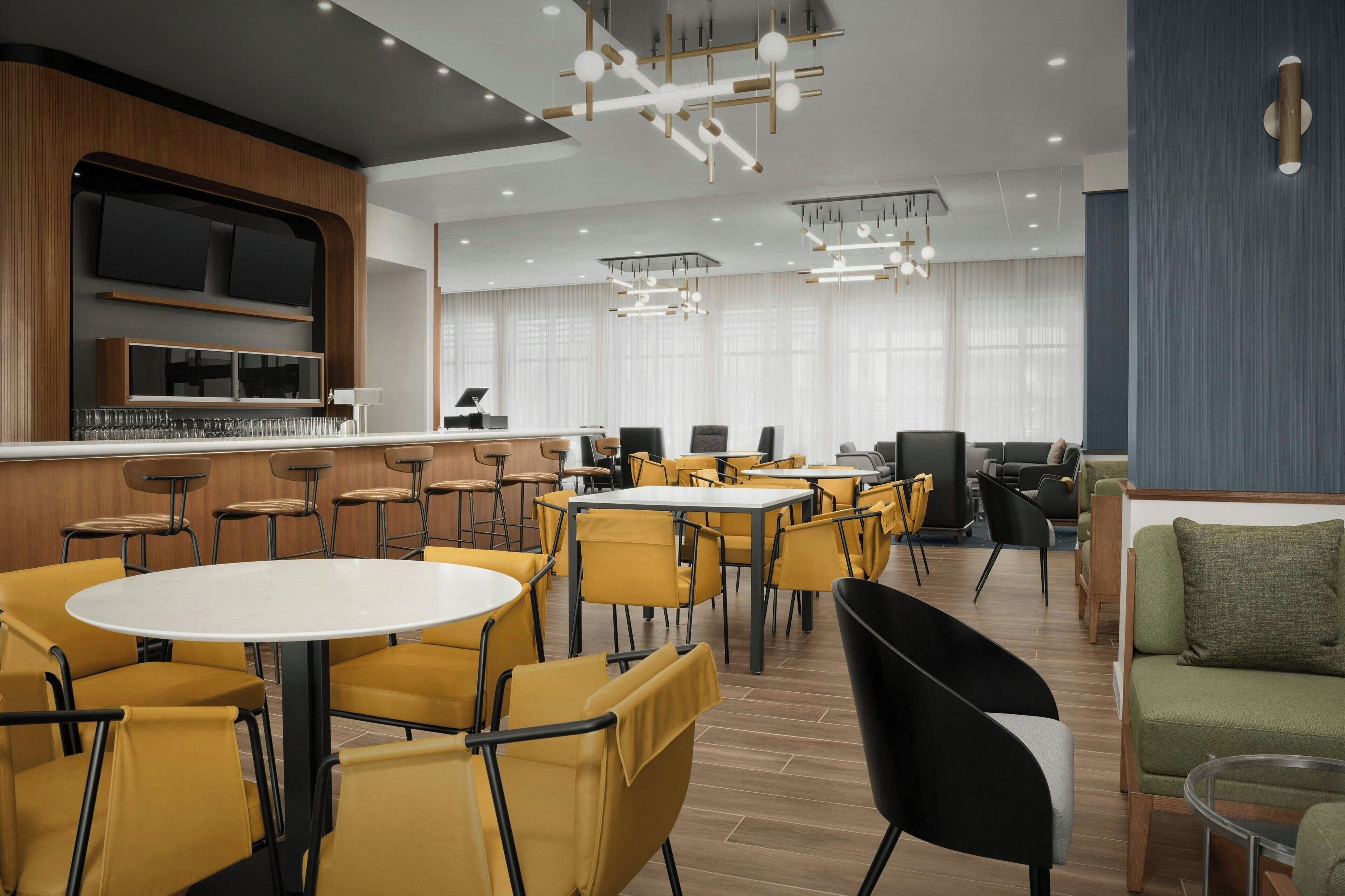 Eine moderne Hotelhalle mit runden Tischen, gelben Stühlen, einer Bar und einer Sitzecke mit Sofas.