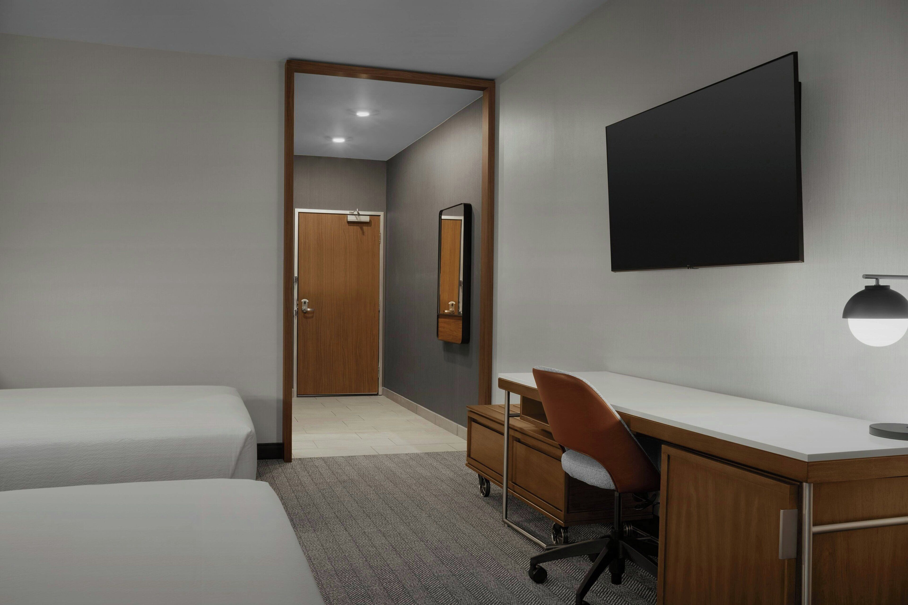 A hotel room with two beds, a desk, a chair, a flat-screen TV, a mirror, and a small dresser.