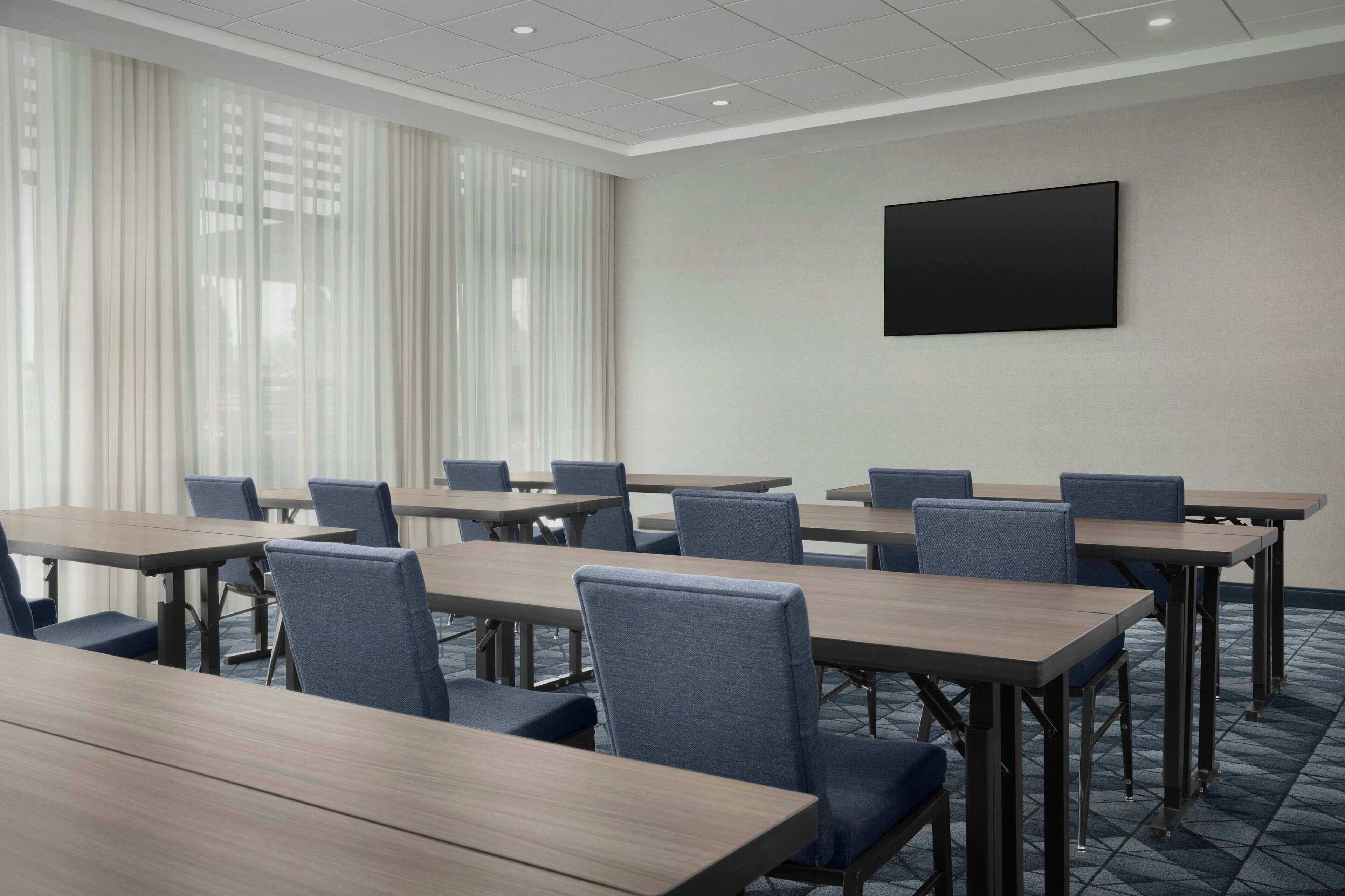 Une salle de conférence dotée de plusieurs tables et de chaises, de grandes fenêtres ornées de rideaux légers et d’un téléviseur à écran plat fixé au mur.