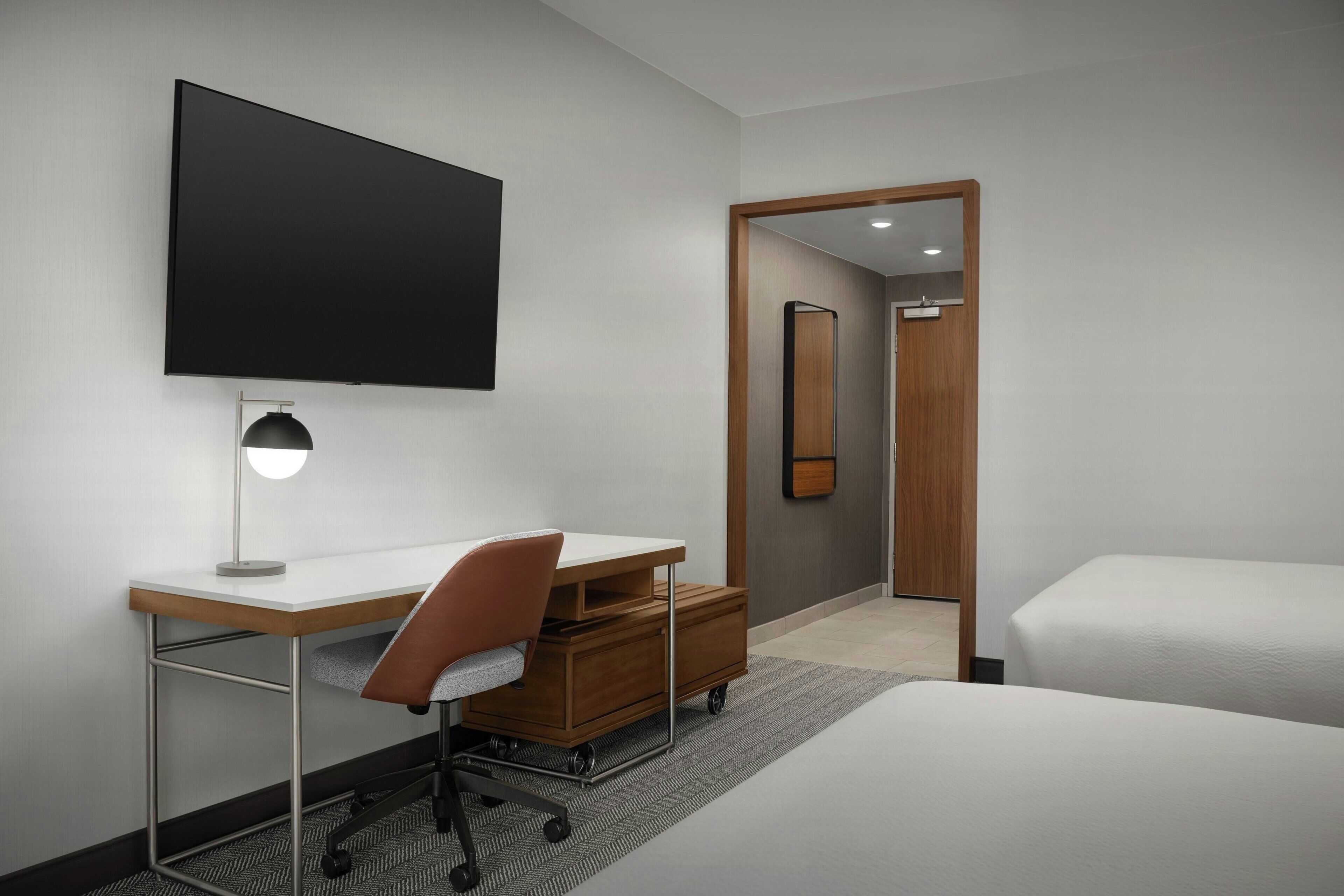 A modern hotel room with a flat-screen TV, a desk with a chair, a bed, and a large mirror.